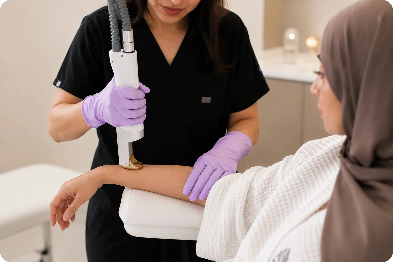 A practitioner in dark scrubs and purple gloves passing a laser handpiece over a client's forearm.